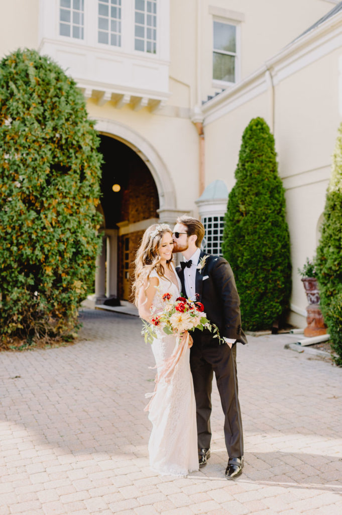 The groom, wearing sunglasses, kisses the bride's temple in a romantic and tender gesture. L Hewitt Photography

