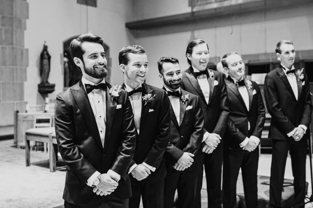 The groom beams with happiness as his bride walks down the aisle, surrounded by his groomsmen. L Hewitt Photography

