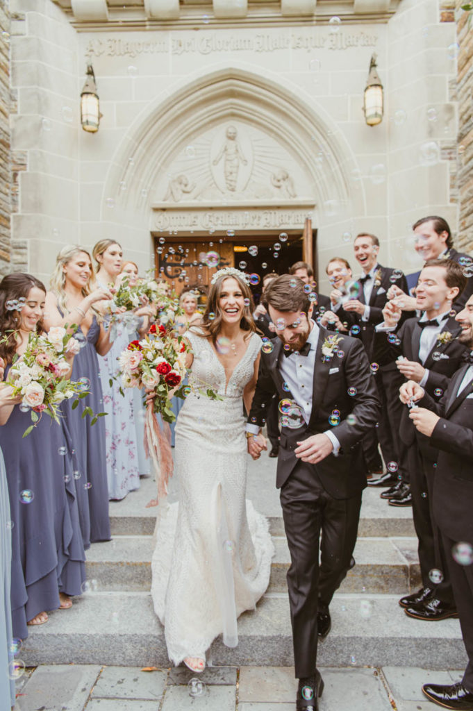 A joyous bride and groom, laughing happily, exit their wedding ceremony surrounded by bubbles and the warmth of their loved ones. L Hewitt Photography

