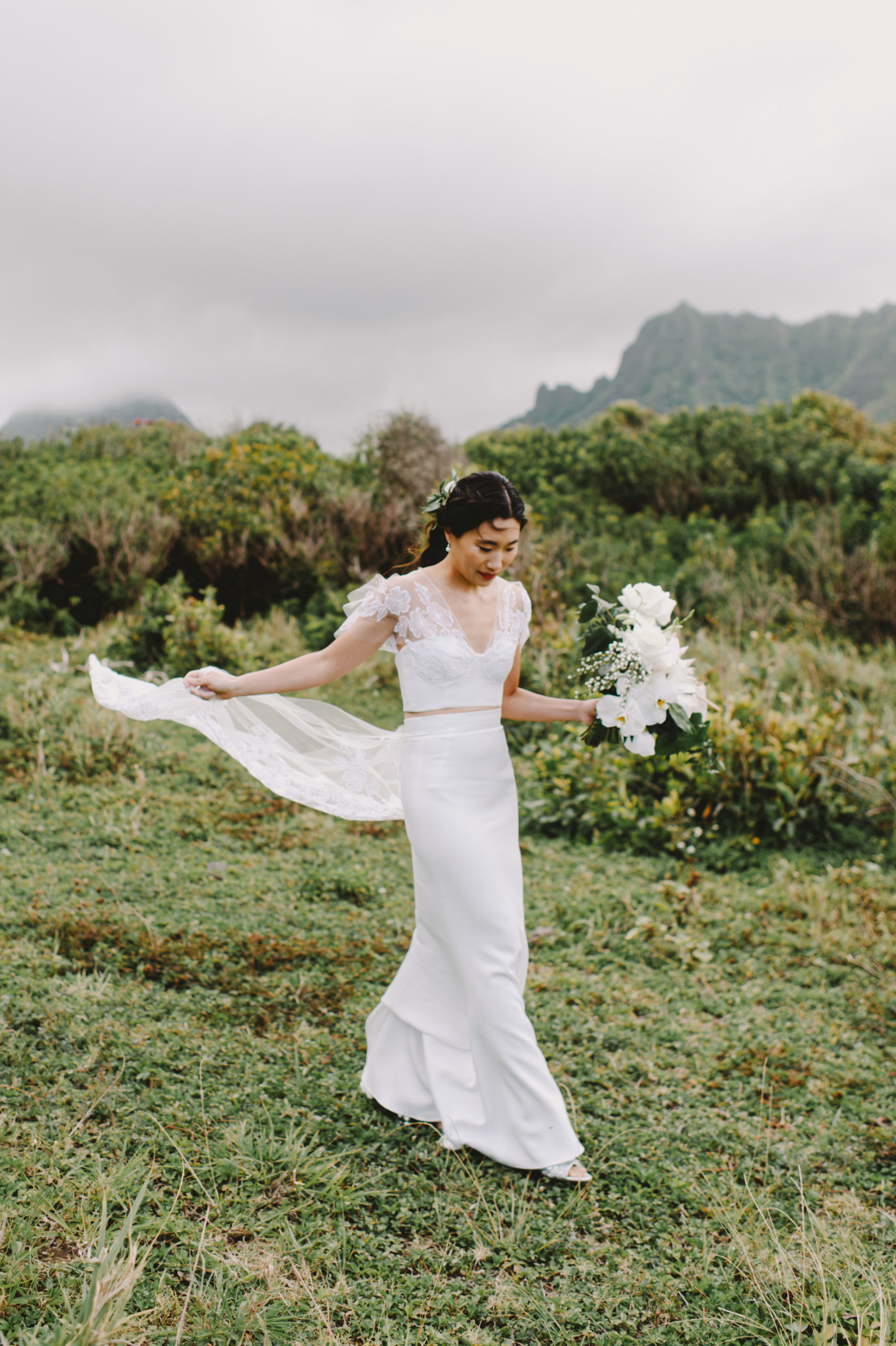 Beautiful bride walking down a verdant hill, bouquet in hand, against the backdrop of Kualoa Ranch's natural beauty L Hewitt Photography
