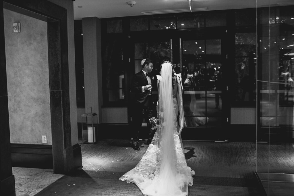 Bride from back has long veil as groom holds door for her.