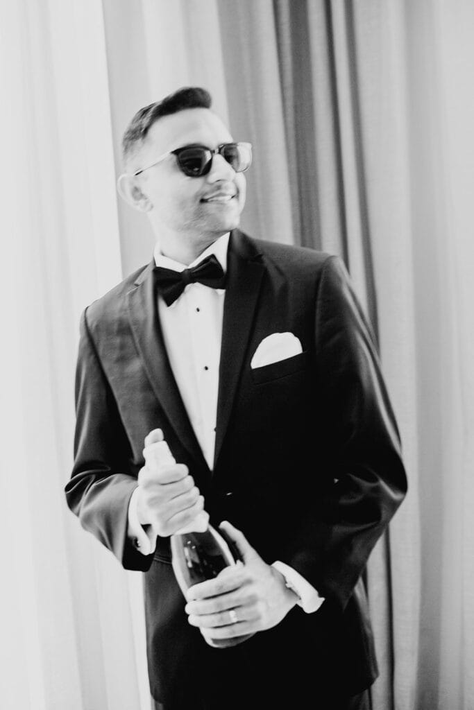 the groom wearing a classic tuxedo with sunglasses and champagne for his Indian wedding reception at Capital One Hall. L Hewitt Photography